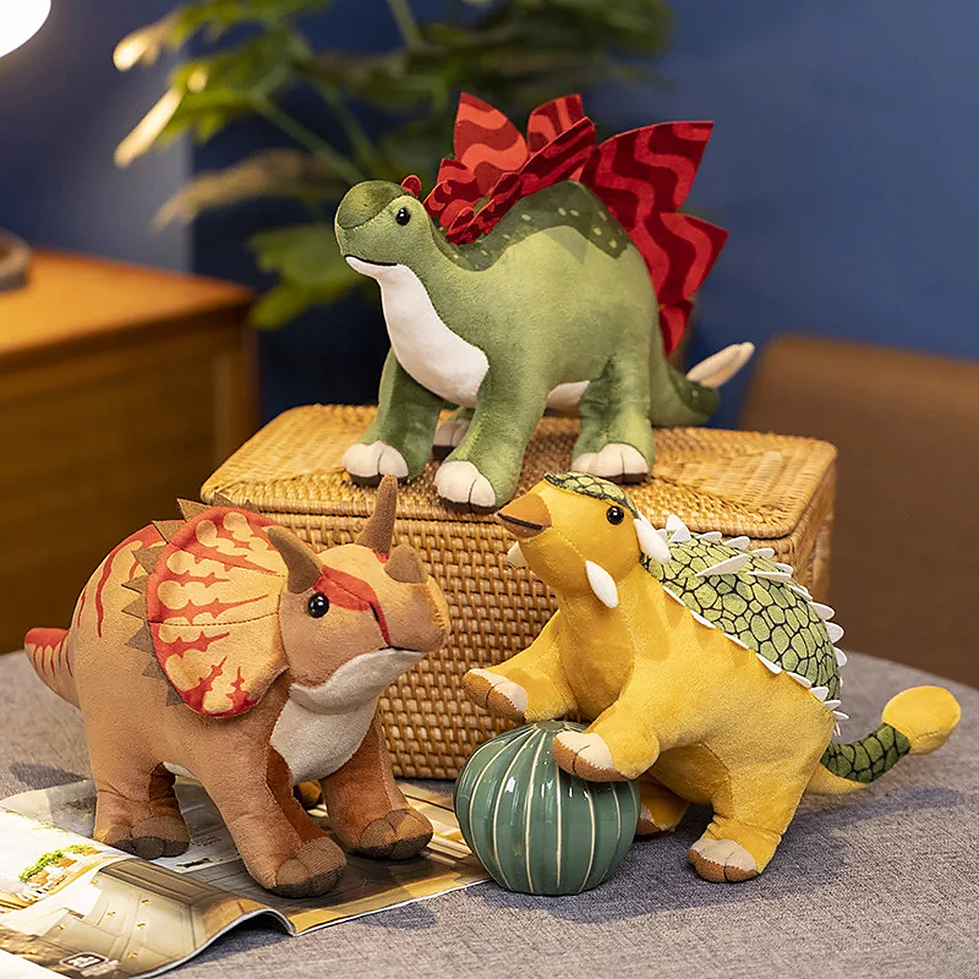Three plush dinosaur toys in various colors and designs, sitting on a table with a decorative item nearby.