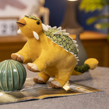 Plush dinosaur toy with yellow body and green scales, sitting beside a decorative item on a magazine.