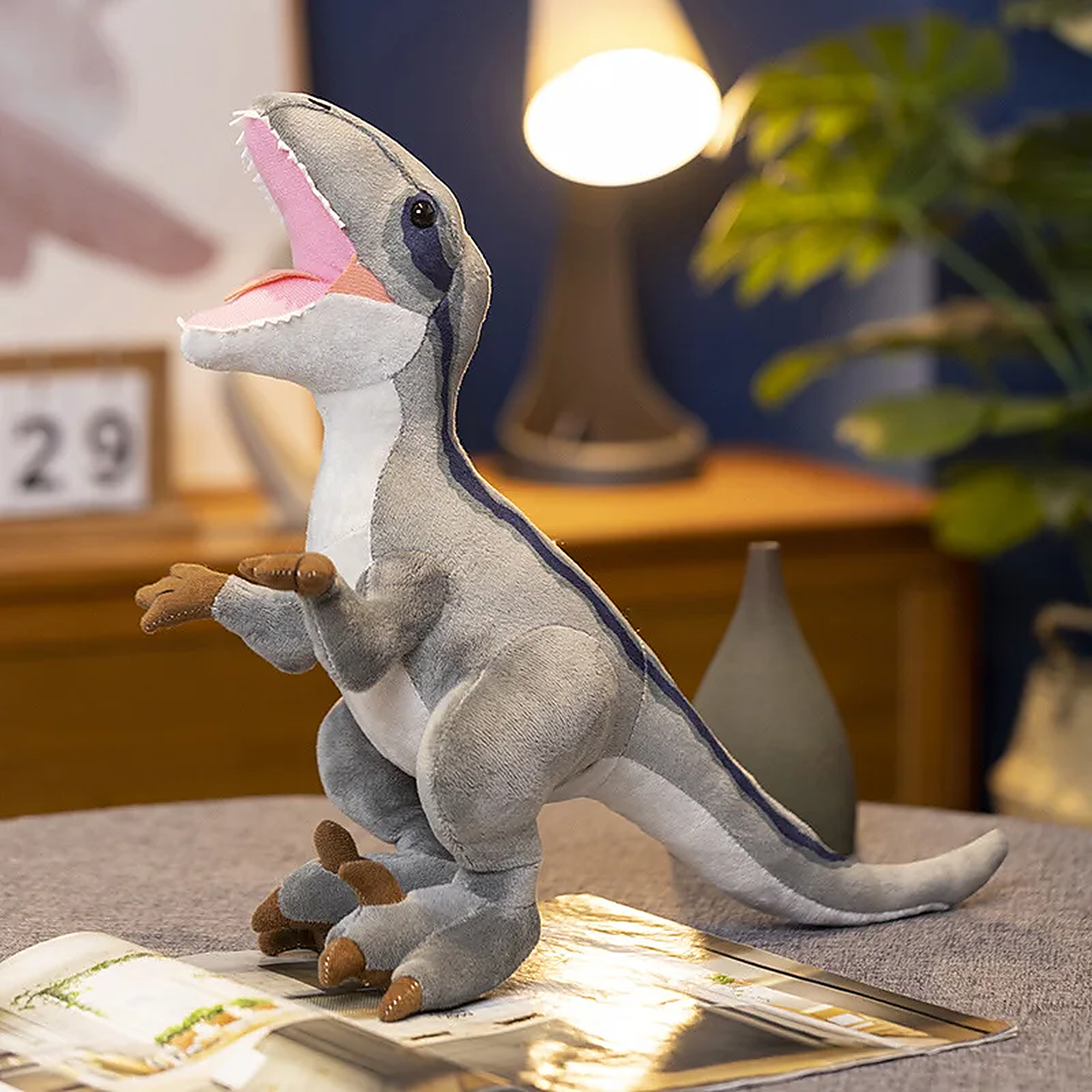 Plush Tyrannosaurus Rex toy with a gray body and pink mouth, sitting on a table with a lamp and plant in the background.