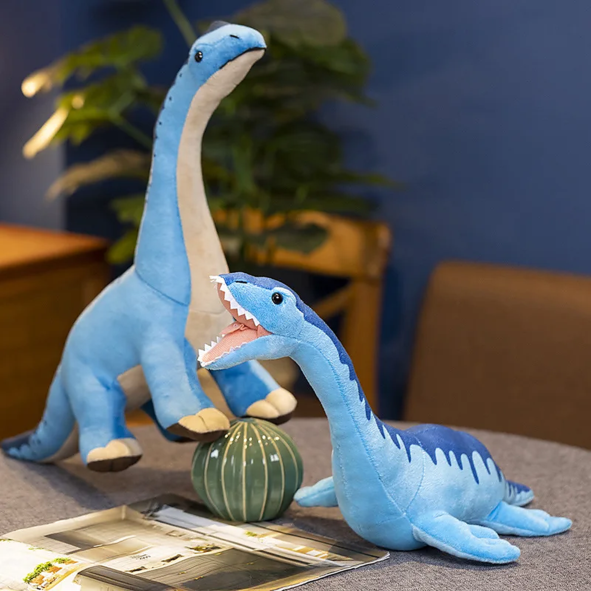 Two blue plush dinosaur toys are displayed on a table with a decorative plant and a magazine.