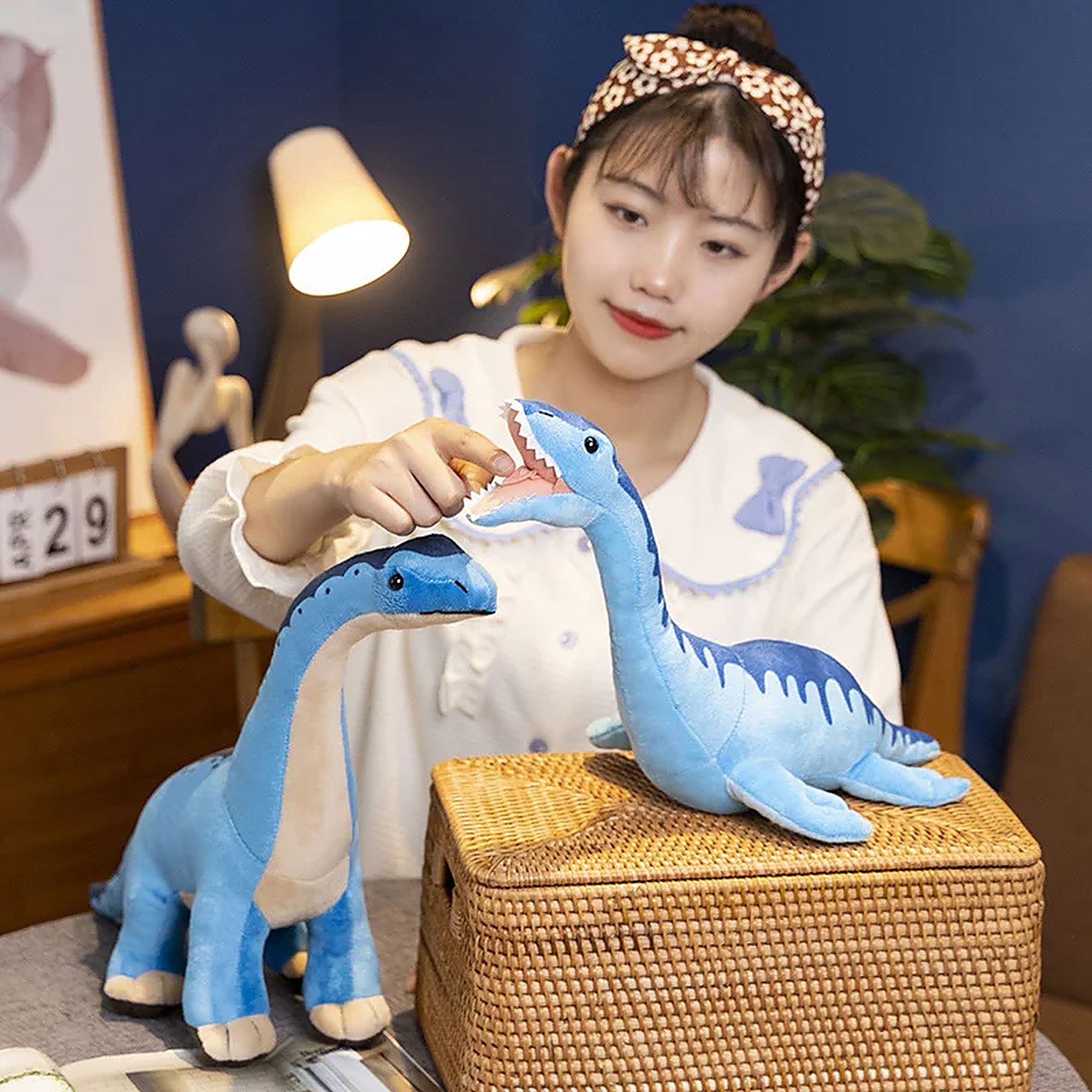 A person interacts with two blue dinosaur plush toys on a table next to a woven basket in a cozy room setting.