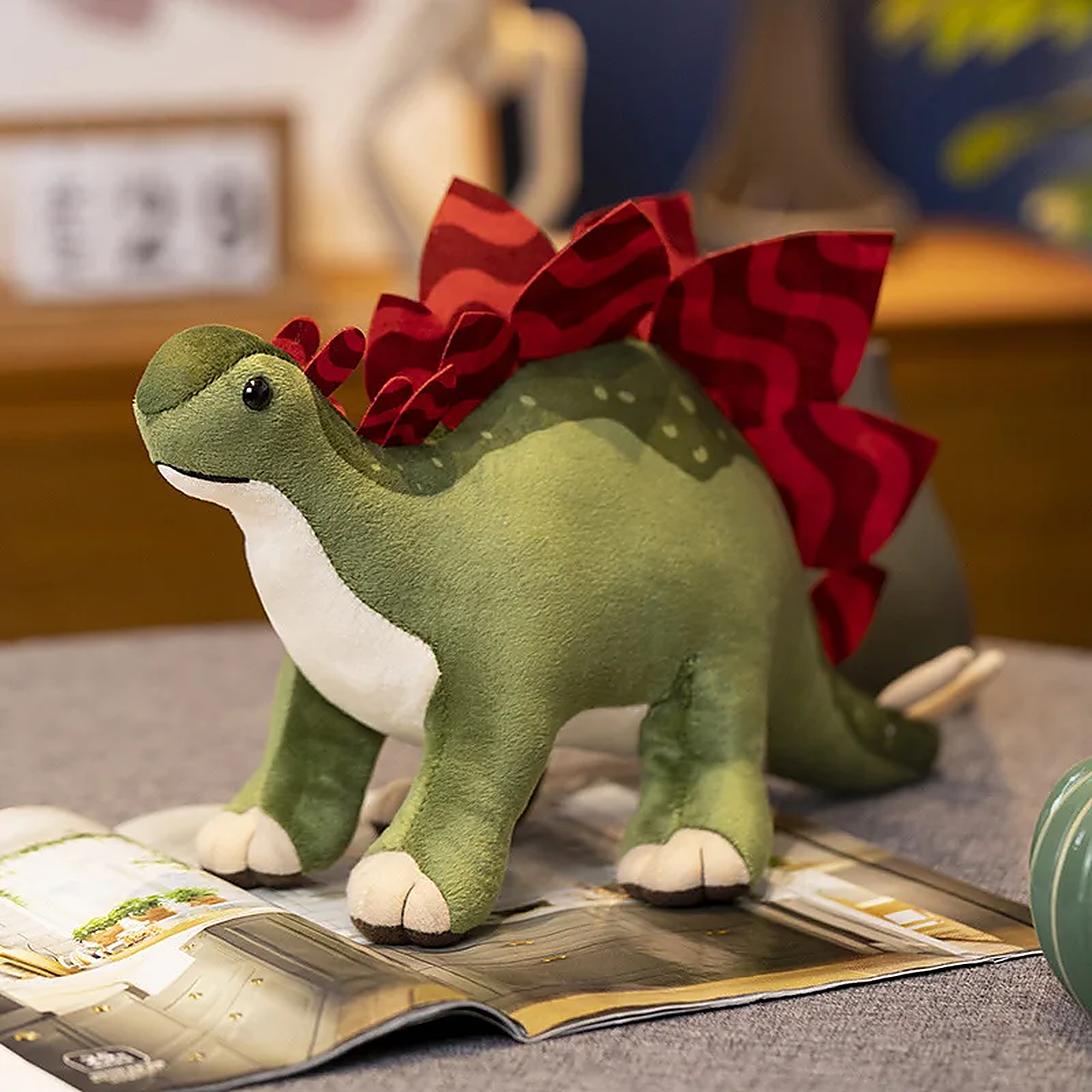 Soft green dinosaur plush toy with a red frill, sitting on a magazine in a cozy room setting.