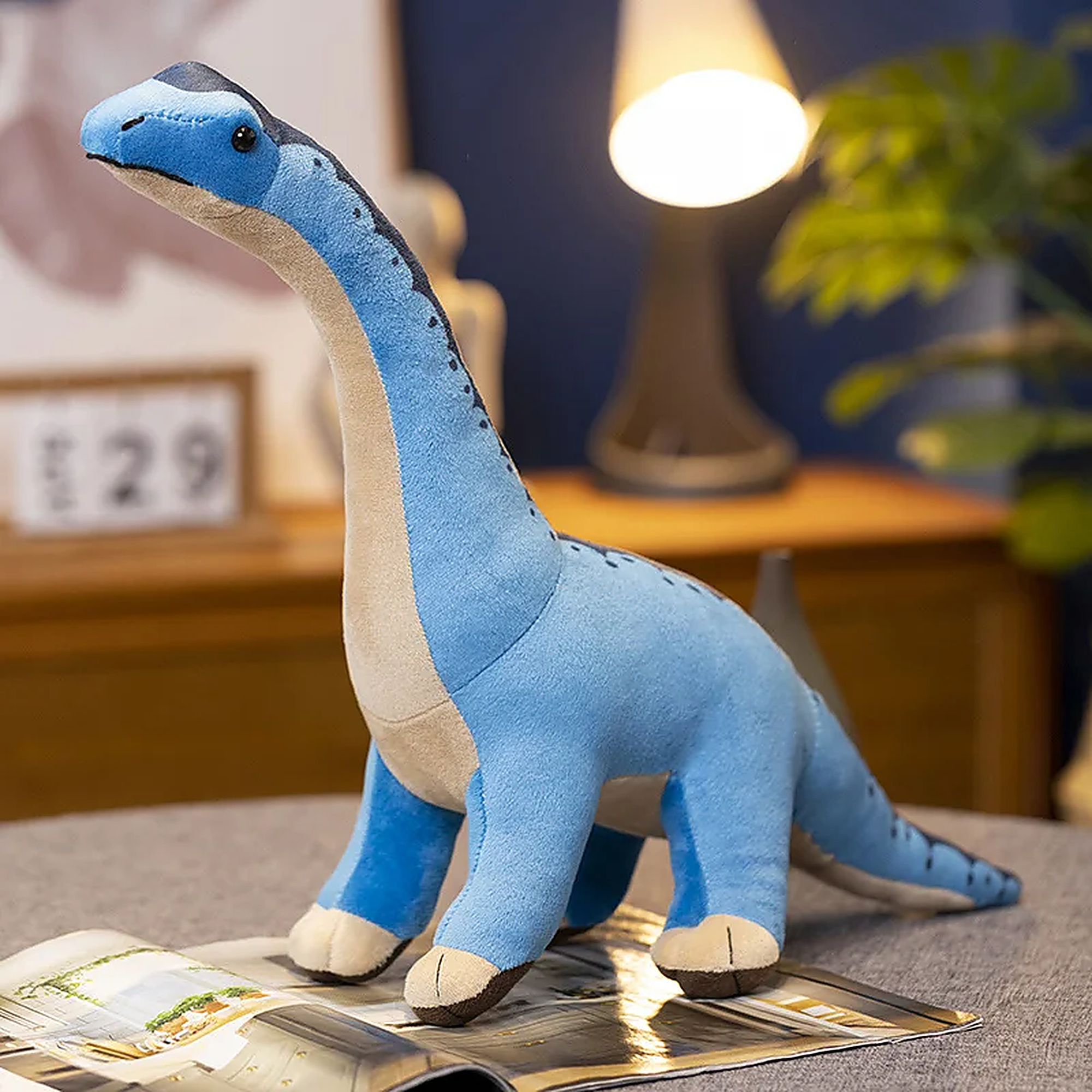 Blue plush dinosaur toy with a long neck and friendly expression, sitting on a table next to a magazine.
