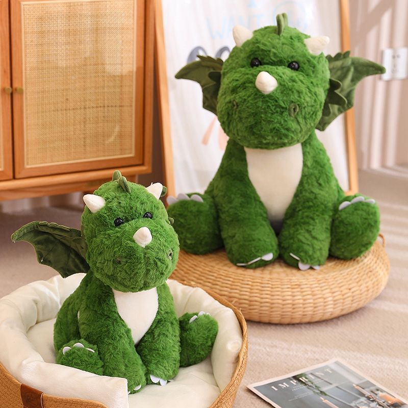 Green plush dinosaur with wings, sitting in a basket, next to a larger version in a cozy setting.