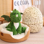 A green plush dinosaur with wings sits in a cozy bed next to a speckled dragon egg-shaped pillow.