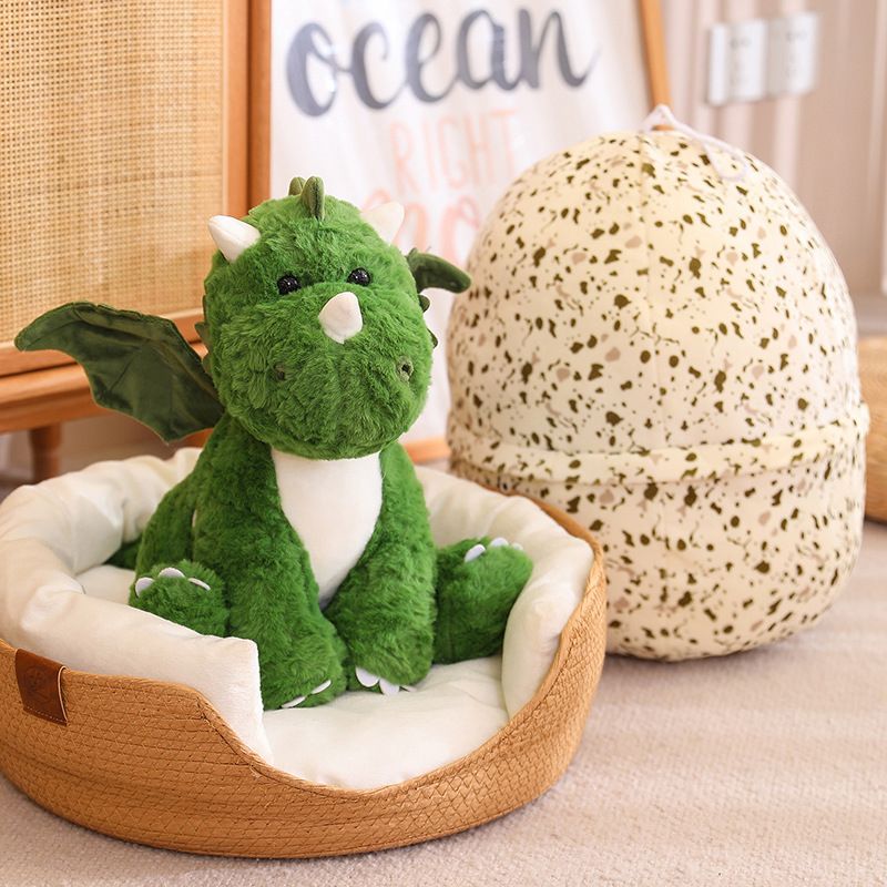 A green plush dinosaur with wings sits in a cozy bed next to a speckled dragon egg-shaped pillow.