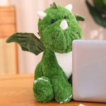 Green plush dinosaur with wings sitting beside a laptop on a wooden table.
