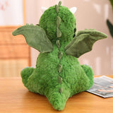 Green plush dinosaur with wings and a textured body, viewed from the back, sitting on a wooden surface.