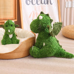 Green dinosaur plush toy sitting next to a smaller version, both with wings, on a soft surface.