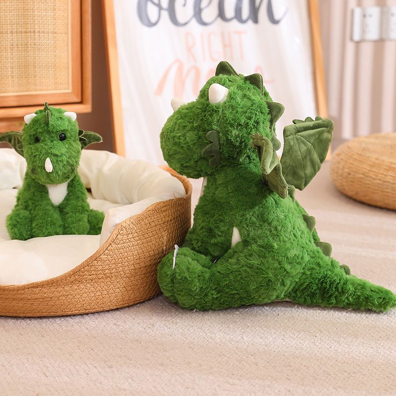 Green dinosaur plush toy sitting next to a smaller version, both with wings, on a soft surface.