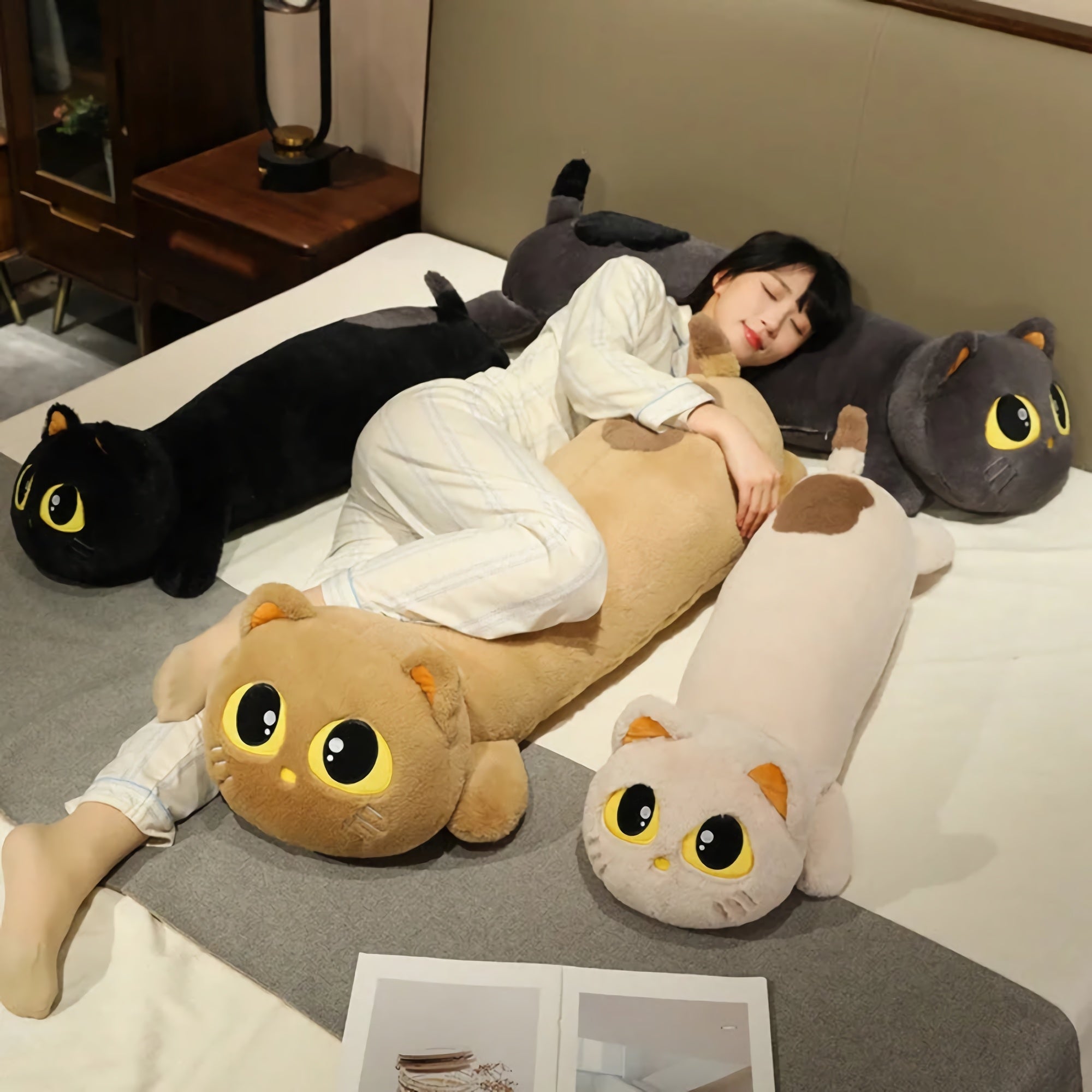 A person in pajamas lies on a bed surrounded by long cat-shaped plush pillows in various colors.