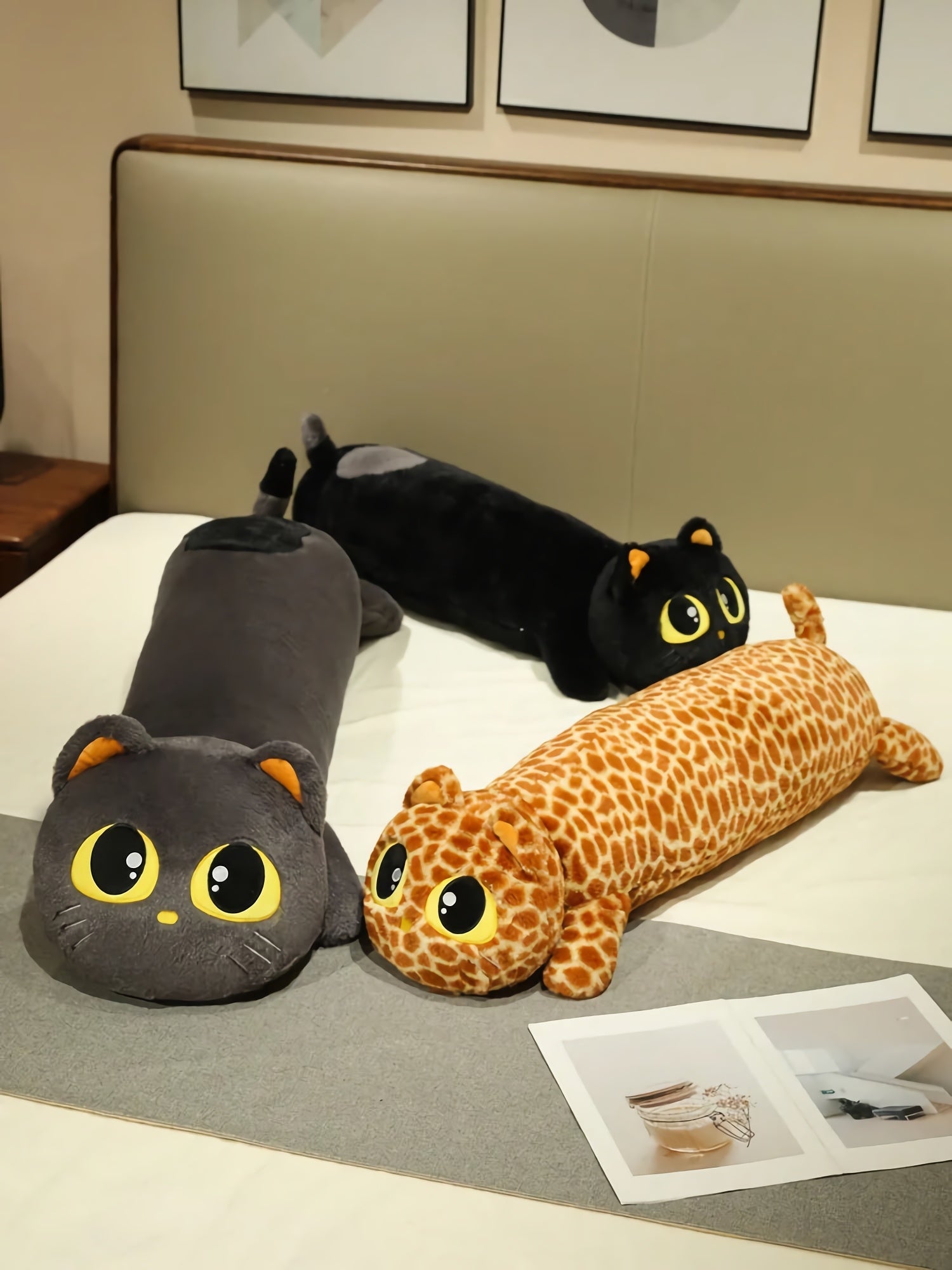 Three long plush pillows shaped like cats and a giraffe, with large eyes, resting on a bed.
