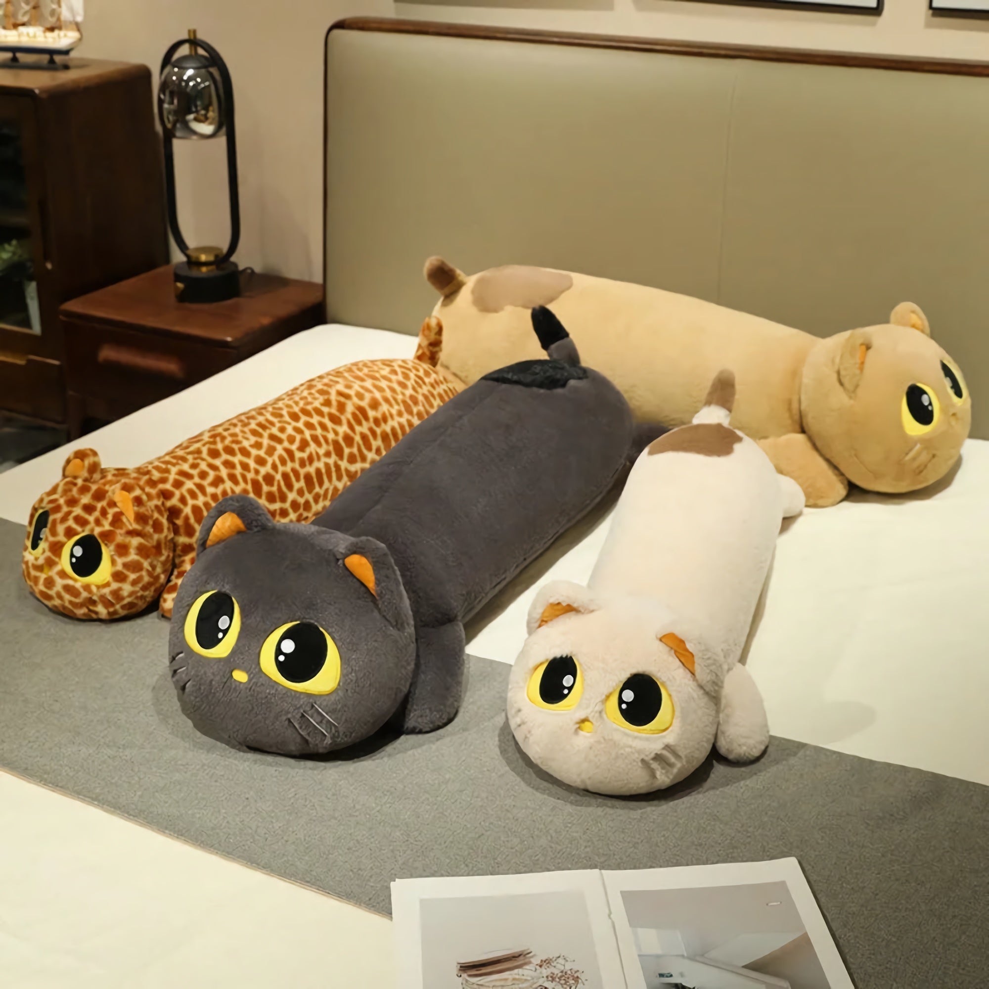 Long plush pillows shaped like cats in various colors and patterns, displayed on a bed with a gray blanket.