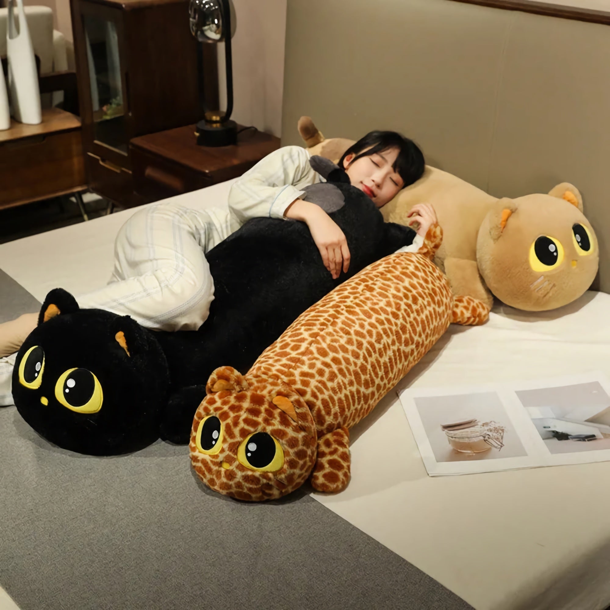 A person sleeps on a bed surrounded by large plush cat and animal pillows in various colors and patterns.