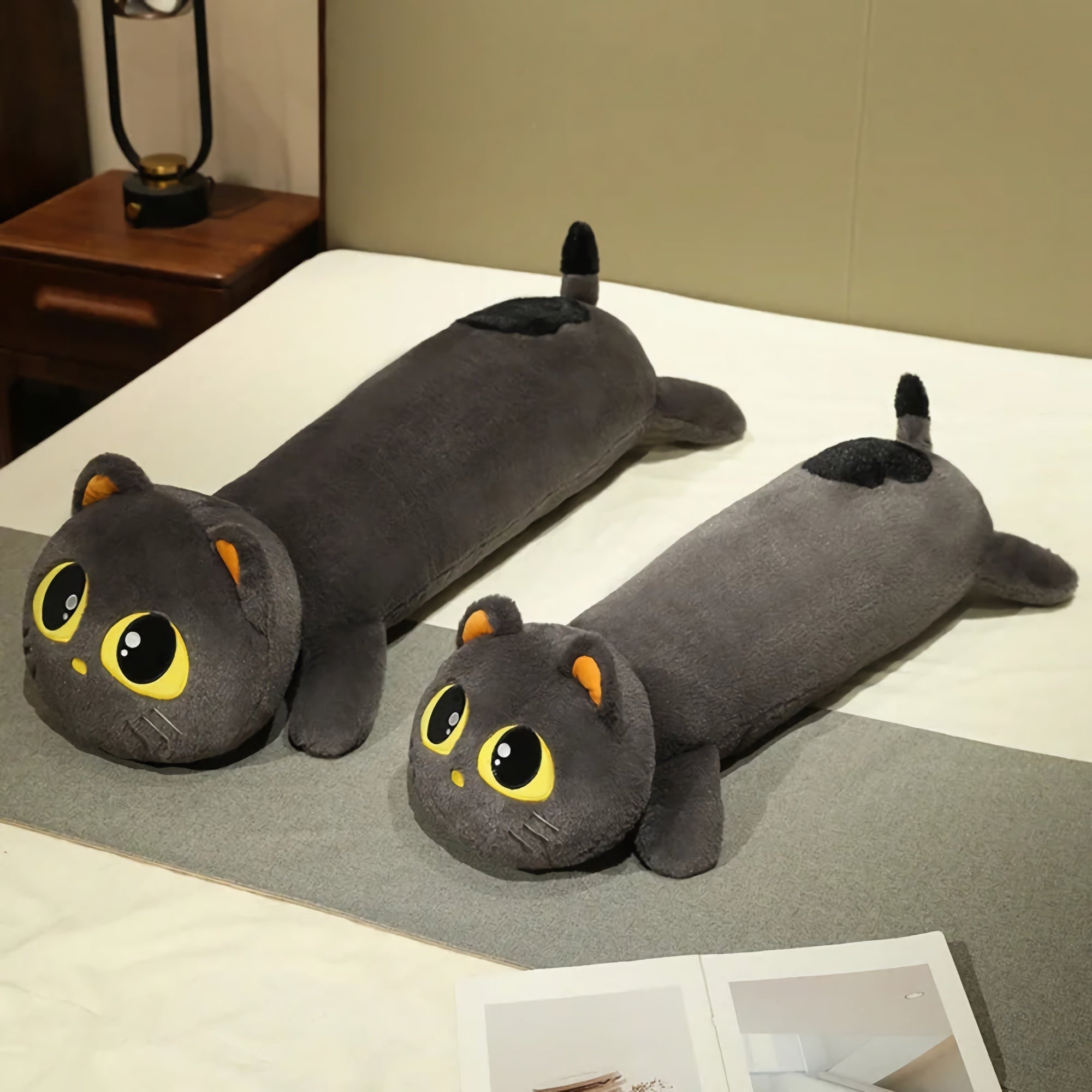 Plush long cat pillow with large eyes, gray fur, and orange ears, resting on a bed with a gray blanket.