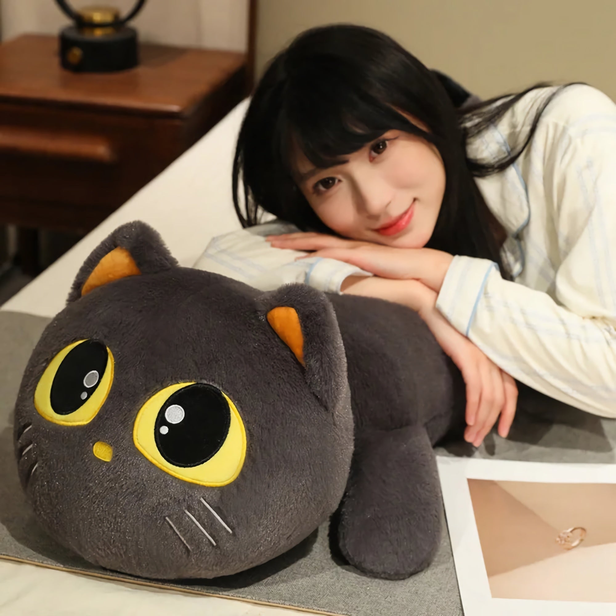A person rests on a bed beside a large, gray cat-shaped plush pillow with big yellow eyes.