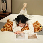 A person in pajamas lies on a bed, reading a book while resting on a long plush giraffe pillow.