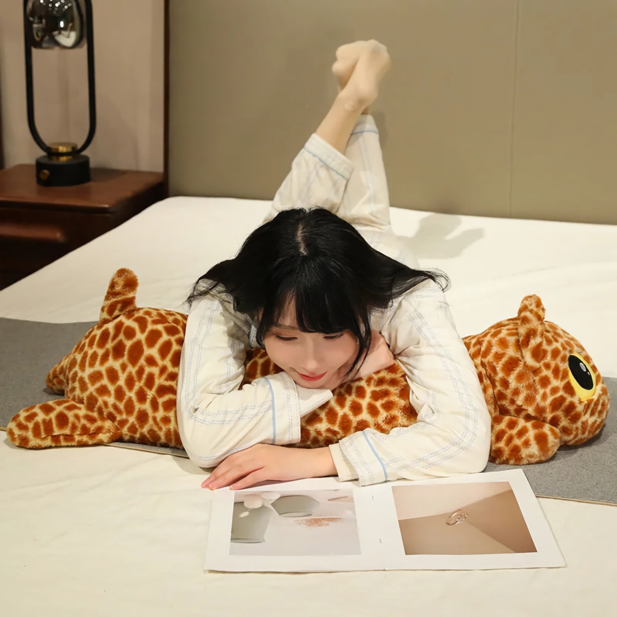 A person in pajamas lies on a bed, reading a book while resting on a long plush giraffe pillow.