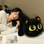 A person rests their head on a long black cat plush pillow with large eyes, lying on a bed with a book nearby.