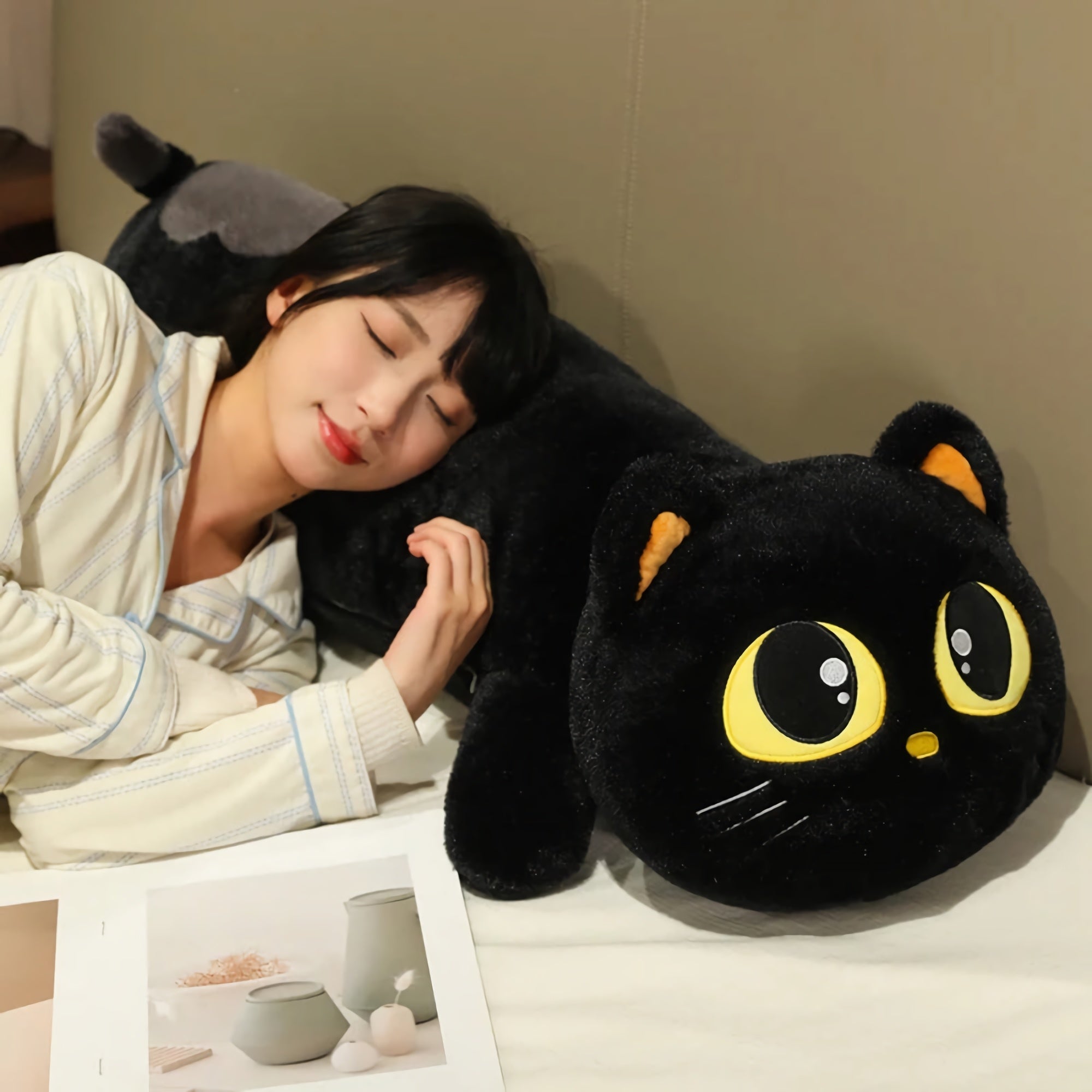 A person rests their head on a long black cat plush pillow with large eyes, lying on a bed with a book nearby.
