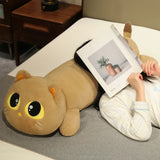 A person lies on a bed with a long cat plush pillow, holding a magazine over their face.