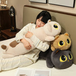 A person in pajamas hugs a long cat-shaped plush pillow, with two smaller plush cats beside them.