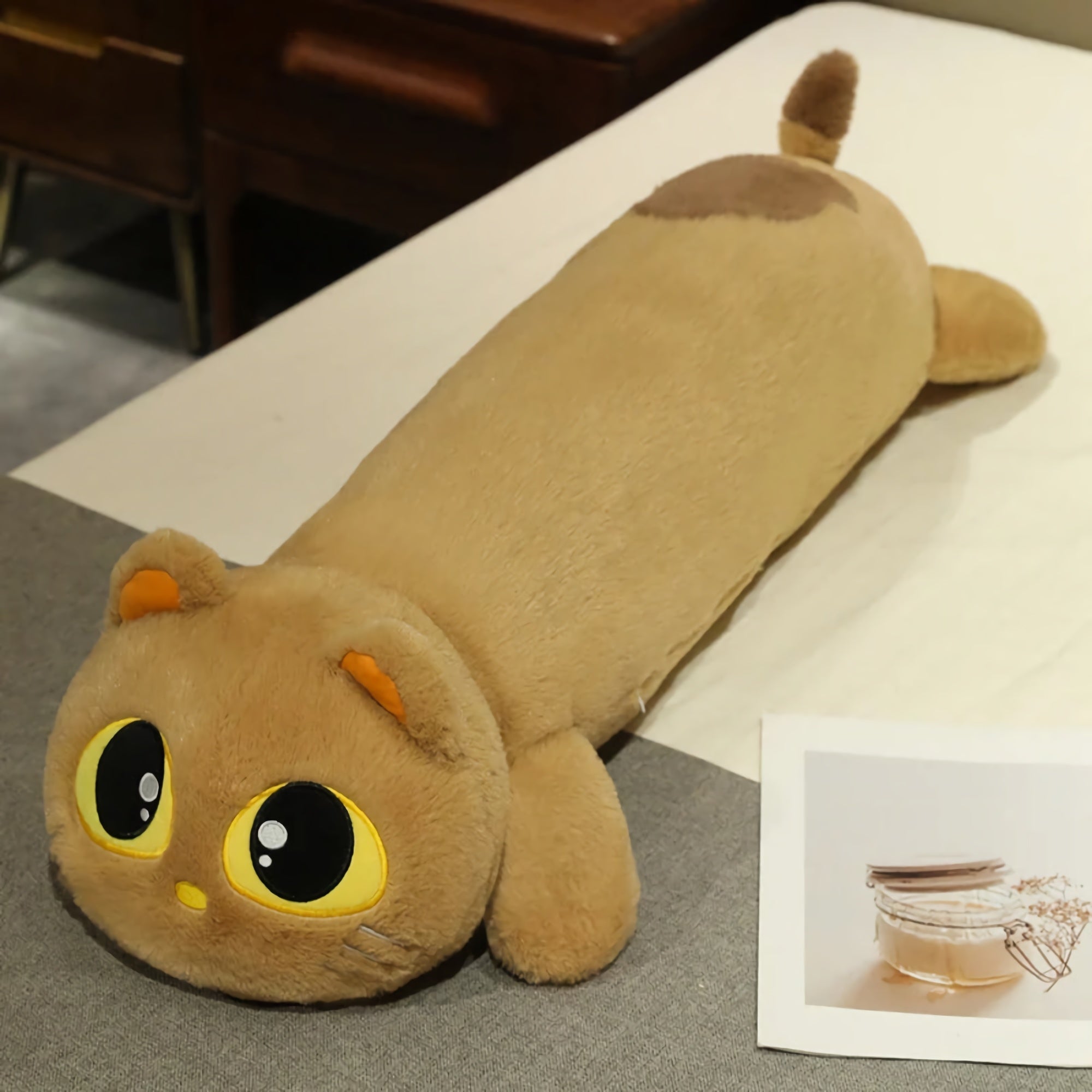 Long plush cat pillow with large eyes, orange ears, and a tail, resting on a bed beside a photo.