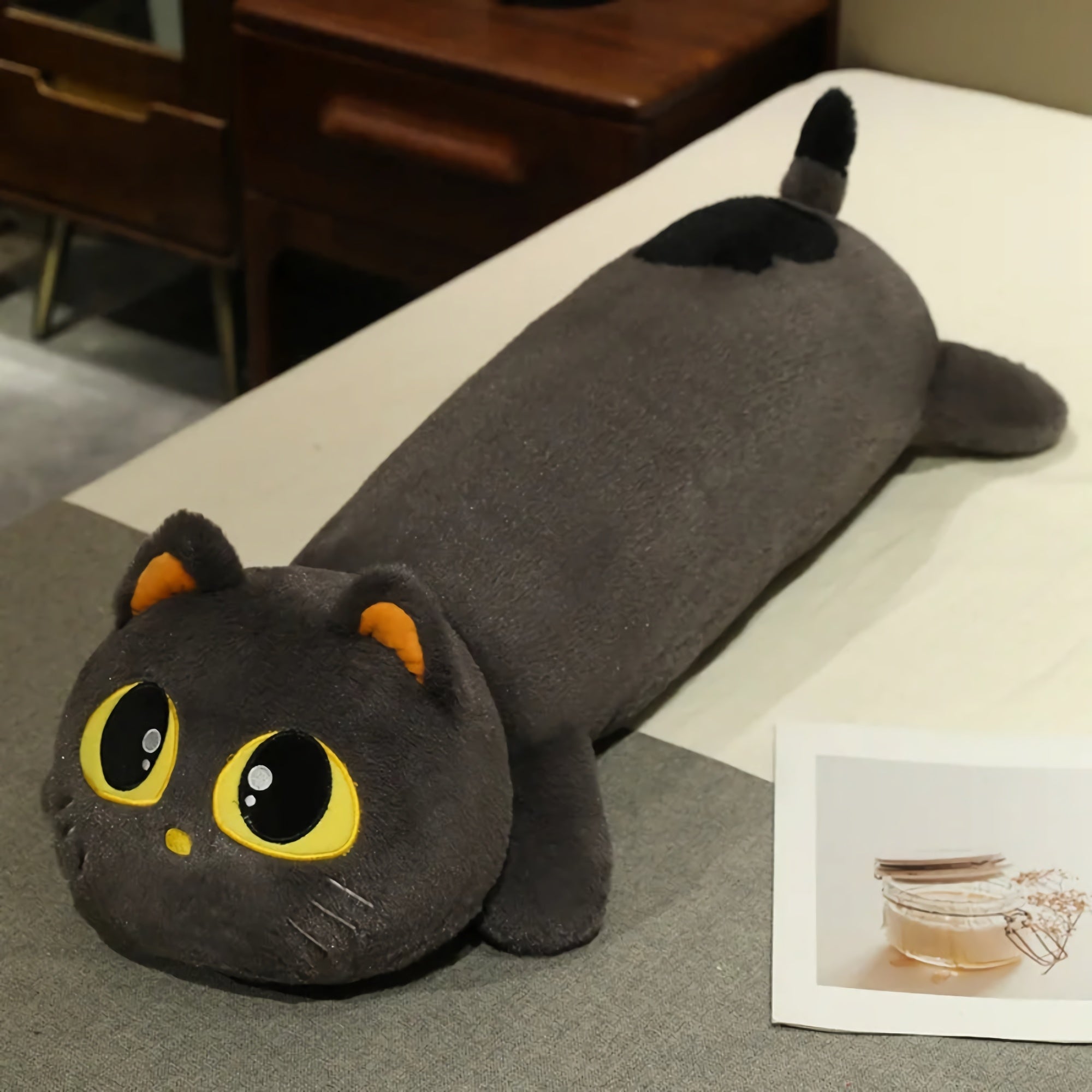 Long gray cat-shaped plush pillow with large yellow eyes, resting on a light surface next to a photo.