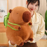 A person holds a large, brown capybara plush with a green backpack, smiling in a cozy setting.