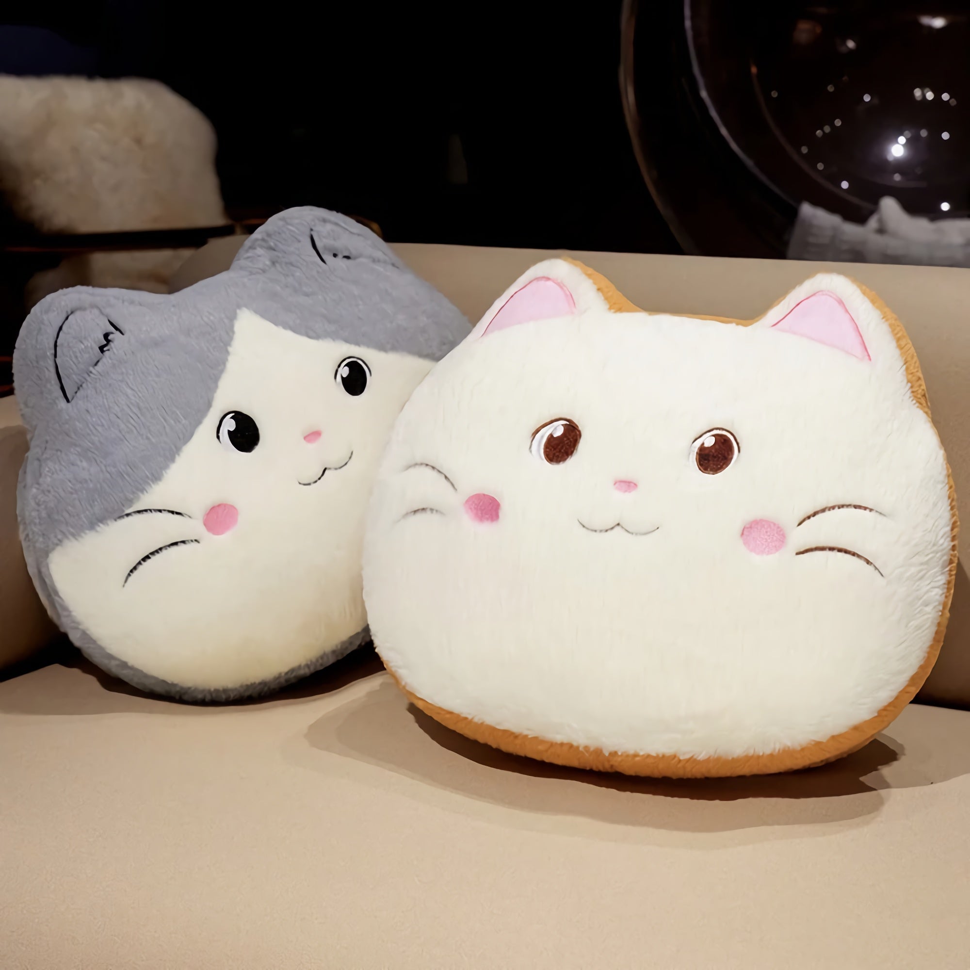 Two plush cat-shaped pillows, one gray and white, the other cream and orange, resting on a beige surface.