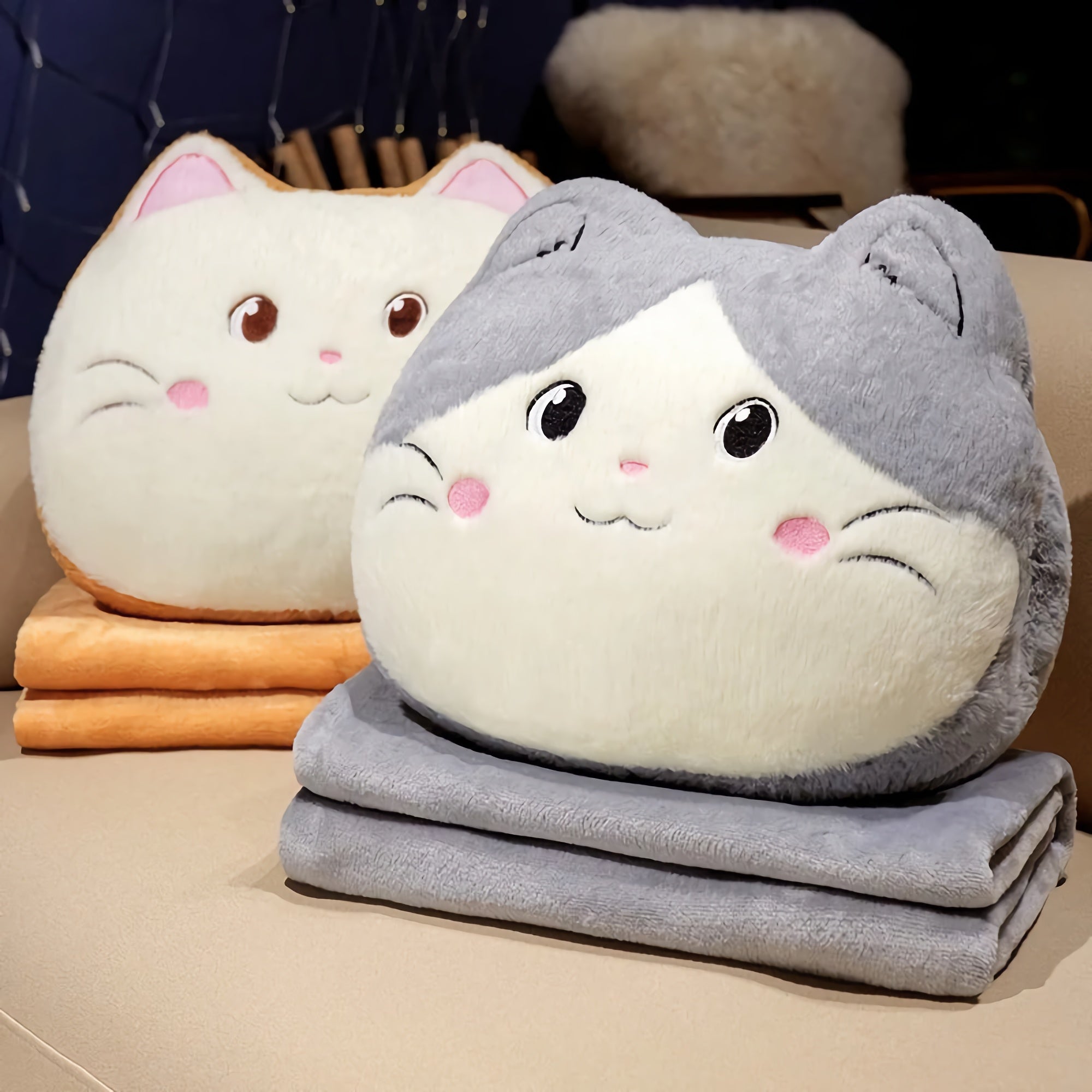 Two plush cat-shaped pillows in gray and cream, resting on folded blankets in orange and gray.