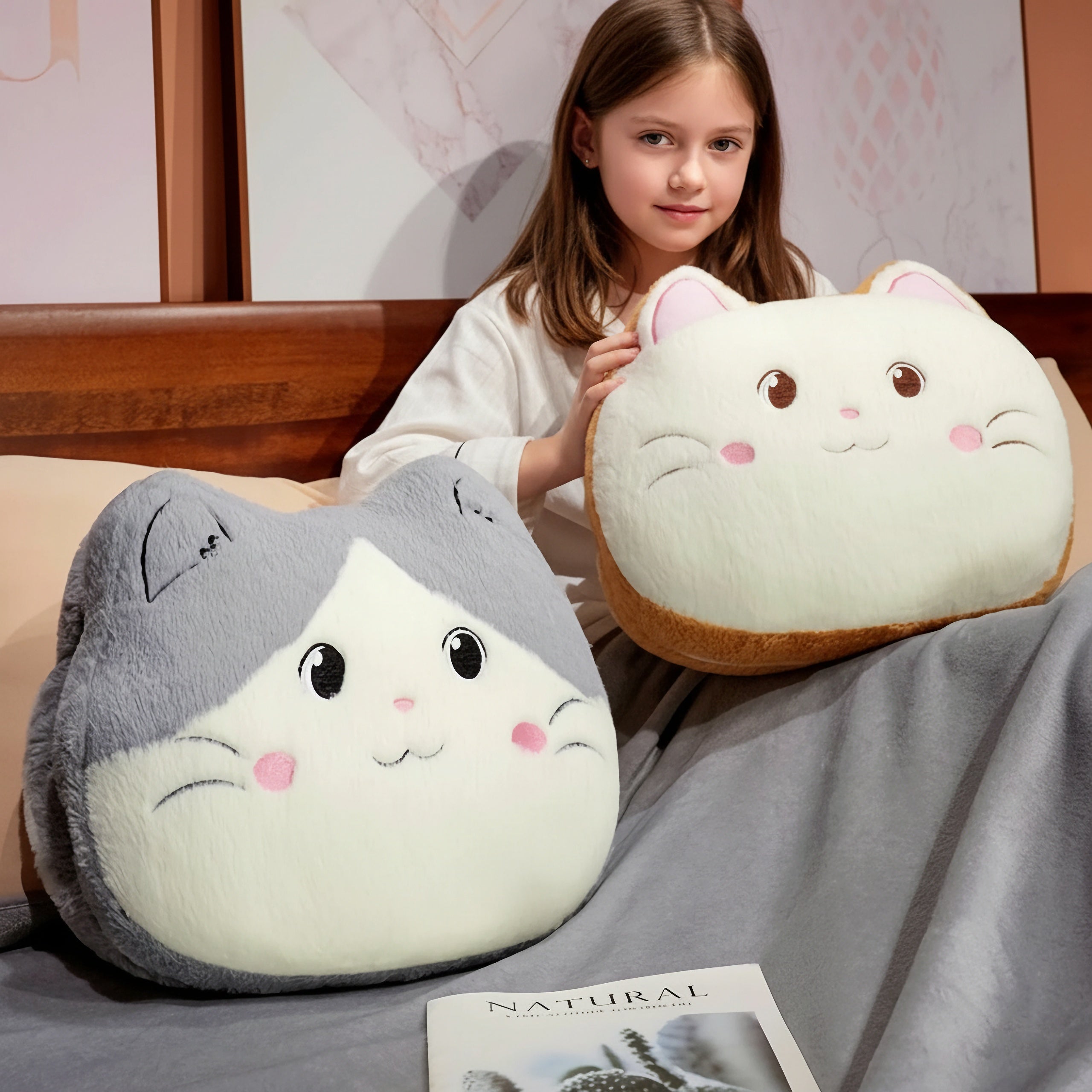 A girl sits on a bed holding two plush cat-shaped pillows, with a soft blanket and a book nearby.