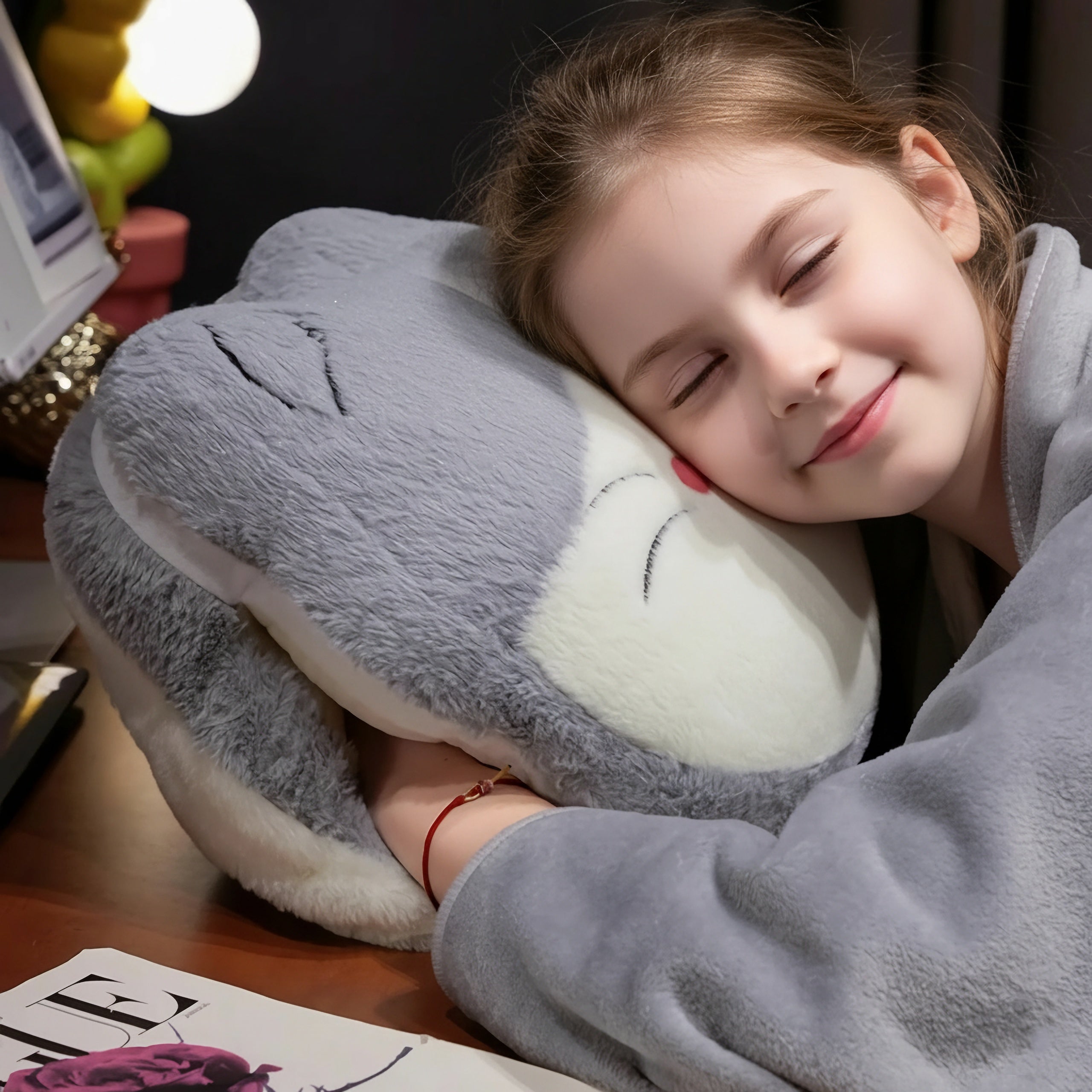 A girl smiles while hugging a plush cat-shaped pillow, wearing a cozy blanket.