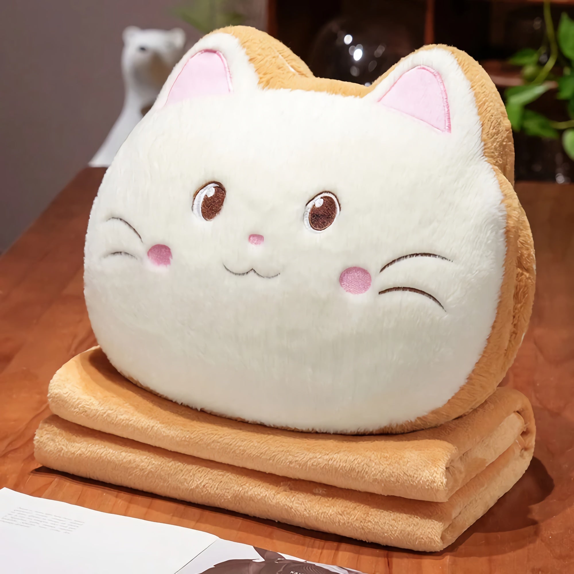 A plush cat-shaped pillow rests on top of a folded blanket on a wooden table.