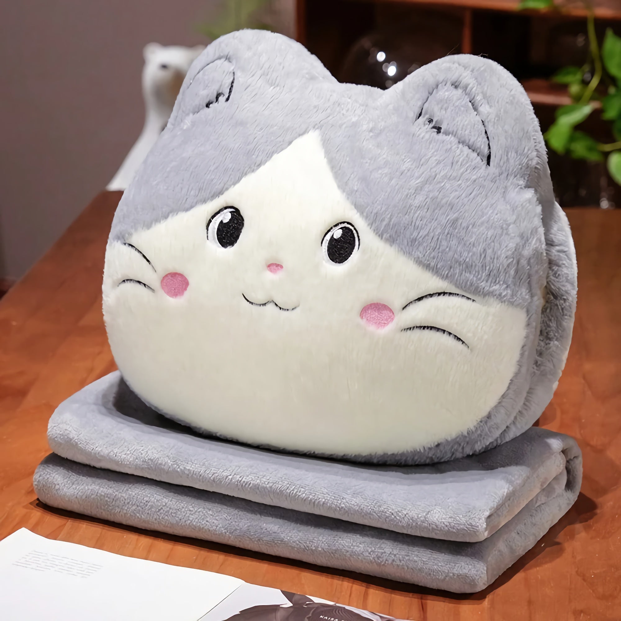 Plush cat-shaped pillow with gray and white fur on a folded gray blanket on a wooden table.