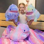 A girl holds two colorful plush dinosaur toys while sitting on a bed with a patterned blanket.