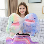 A girl smiles while holding two colorful dinosaur plush toys, sitting on a couch with a patterned blanket.