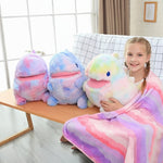 A girl sits on a sofa holding a colorful plush dinosaur pillow, wrapped in a matching blanket, with two similar pillows nearb