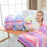 A girl sits on a sofa holding a colorful plush dinosaur pillow, wrapped in a matching blanket, with two similar pillows nearb