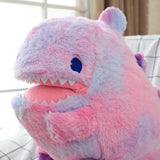 Plush dinosaur pillow in pastel colors with a smiling face and small spikes, resting on a checkered blanket.
