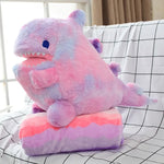 A plush dinosaur pillow in pink and purple sits on a colorful blanket, both on a grid-patterned surface.