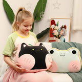 A child holds a plush pillow shaped like a cat, sitting next to a frog-shaped pillow on a cozy blanket.