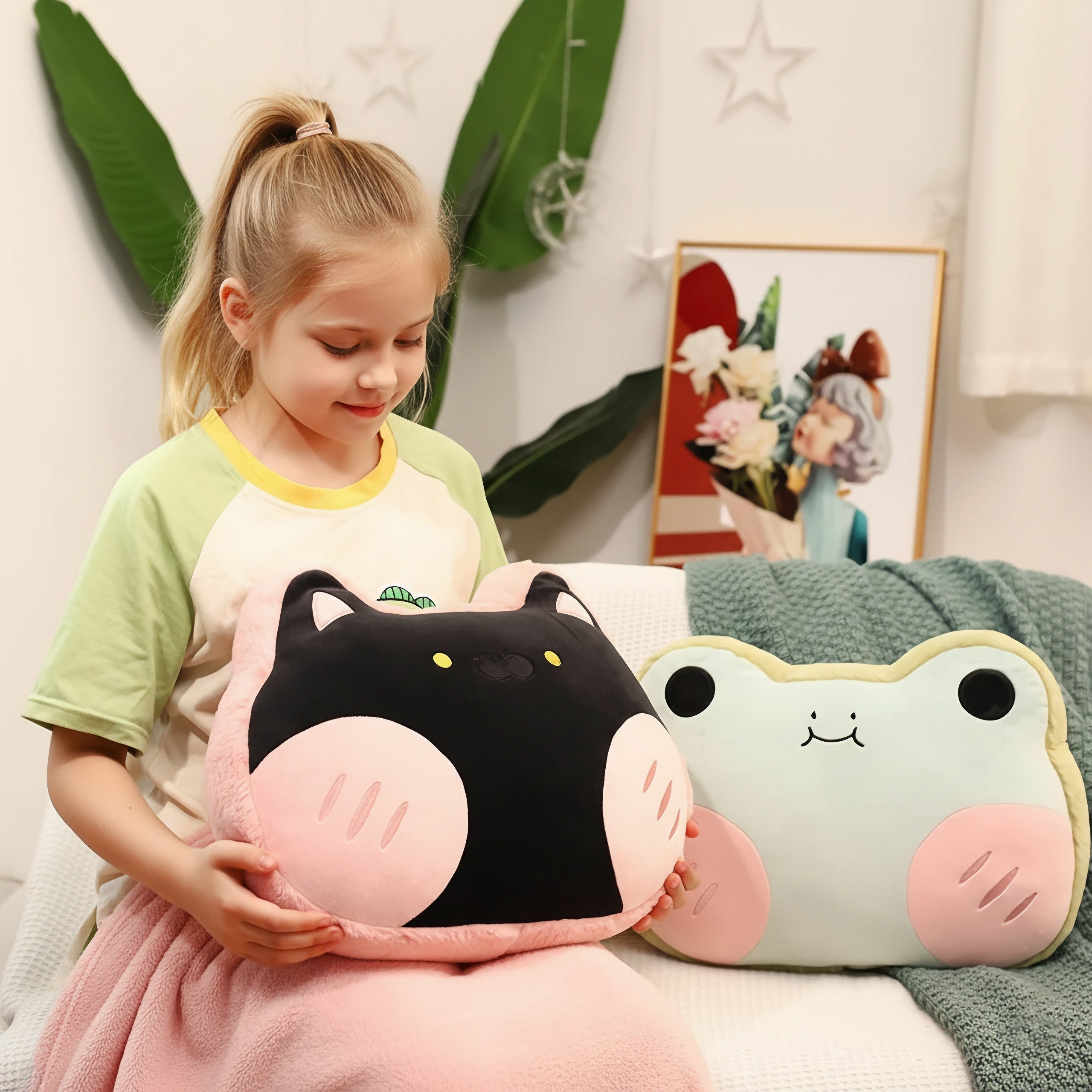 A child holds a plush pillow shaped like a cat, sitting next to a frog-shaped pillow on a cozy blanket.