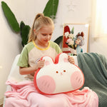 Girl holding a plush pillow shaped like a cute animal face, sitting on a pink blanket.