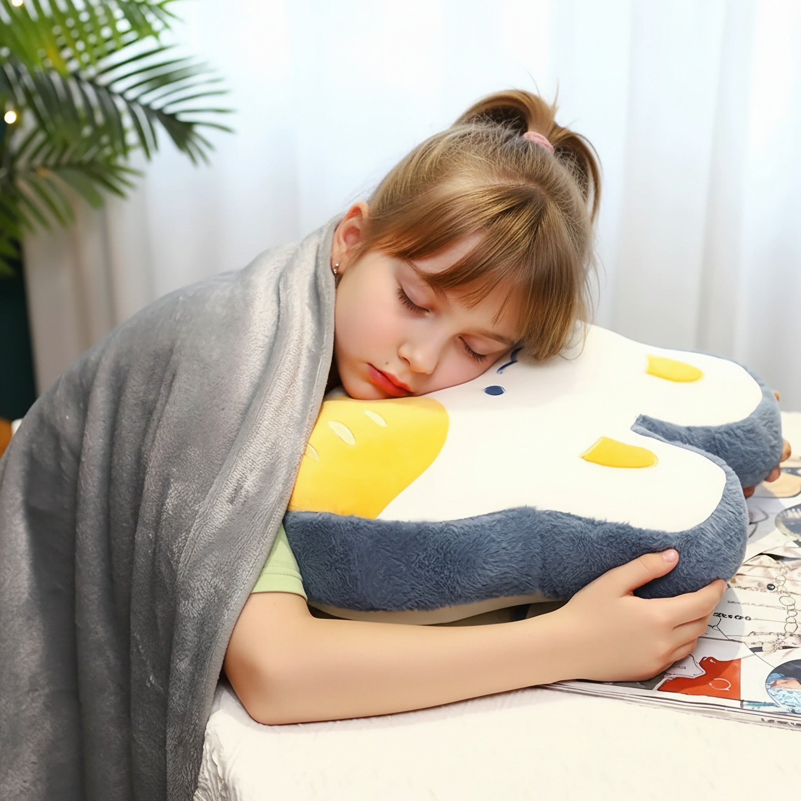 Child sleeping with a gray blanket, hugging a plush pillow shaped like an animal, in a cozy indoor setting.