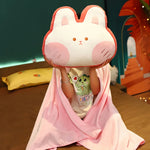 Child sitting on a bed, holding a plush bunny pillow while wrapped in a pink blanket.