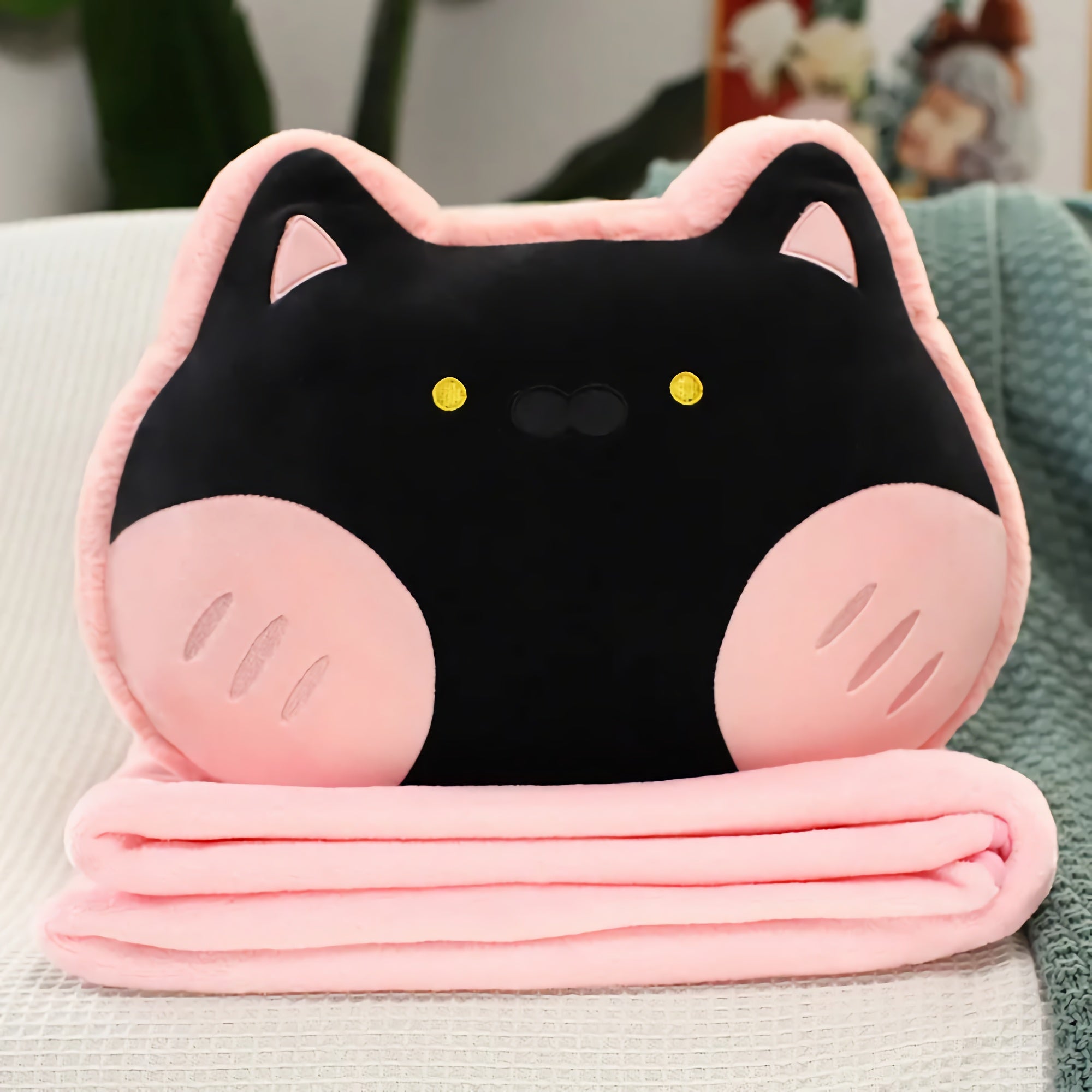 Plush pillow shaped like a cat face with a folded pink blanket underneath.