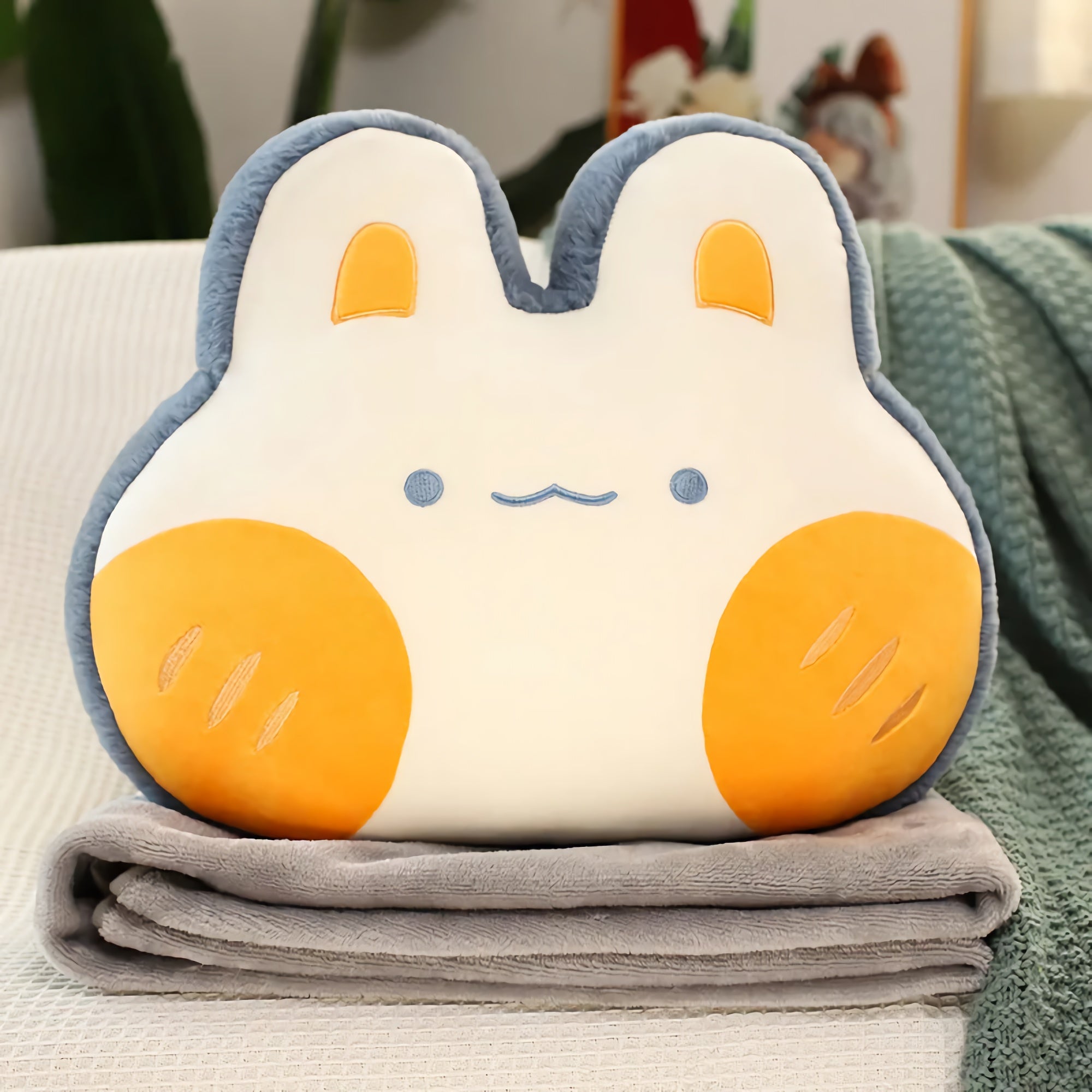 Plush pillow shaped like a bunny with orange cheeks, resting on a folded gray blanket.