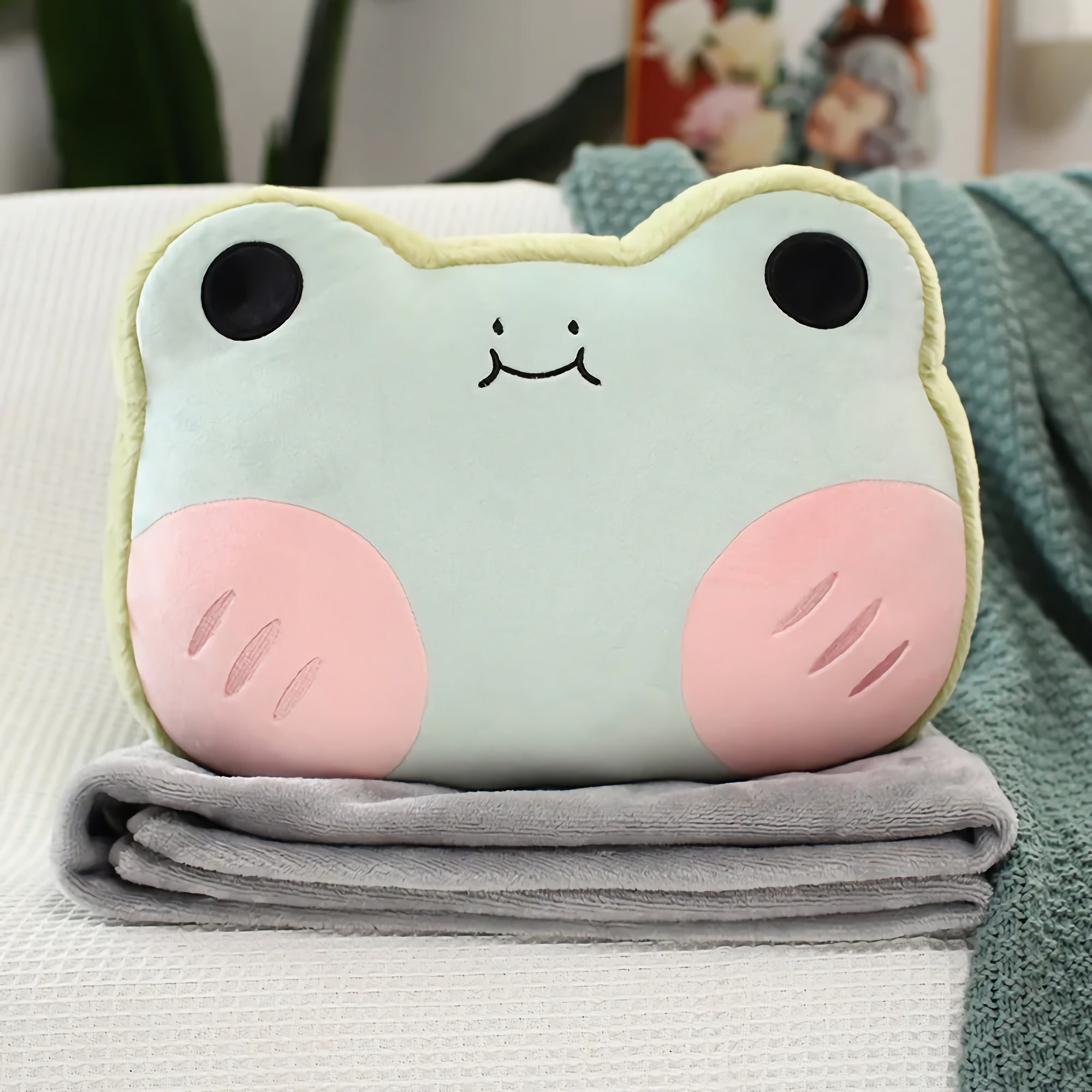 A plush frog pillow with pink cheeks rests on a folded gray blanket.