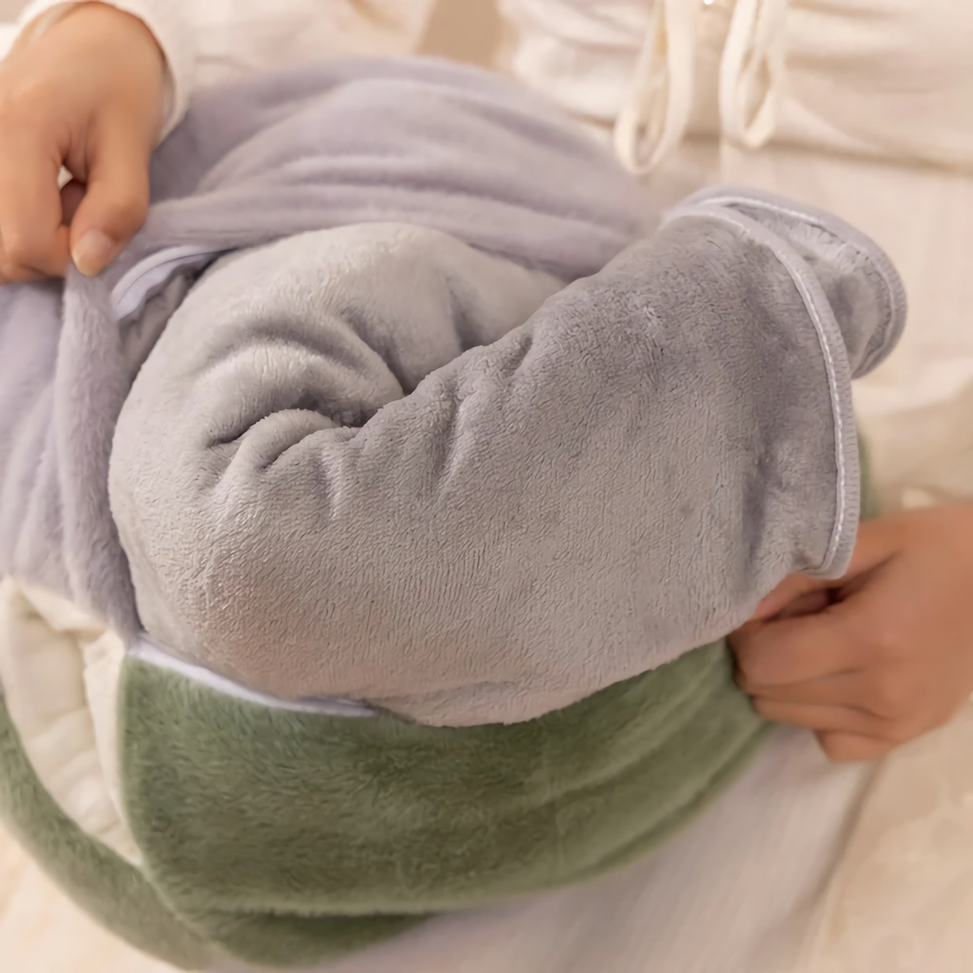 A person holds a rolled-up travel blanket and plush pillow set in soft gray and green fabric.